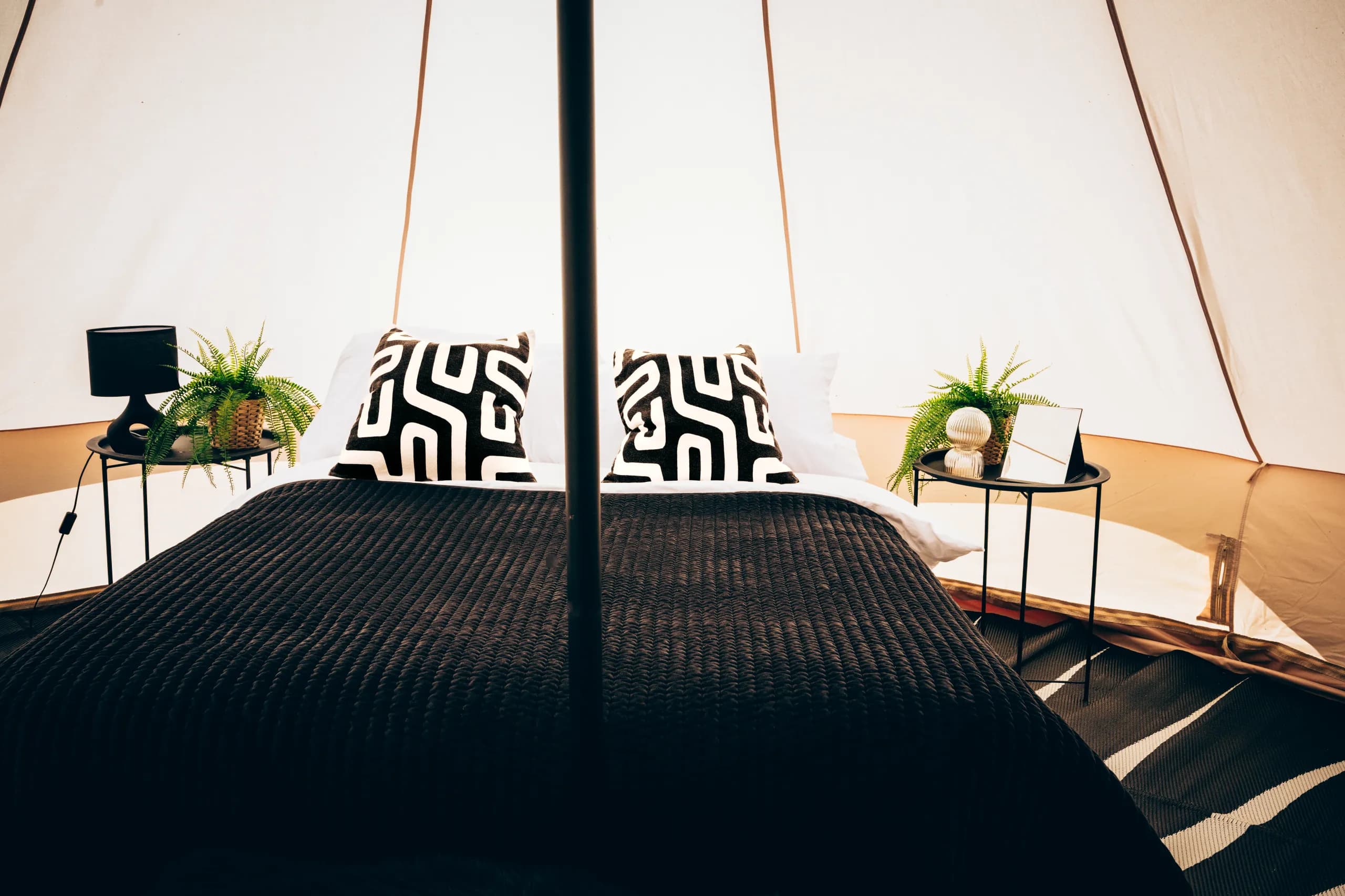 A double bed with a black throw and patterned cushions, flanked by bedside tables with a lamp, ferns and a mirror, inside a Hotel Melody Encore Lounge bell tent.