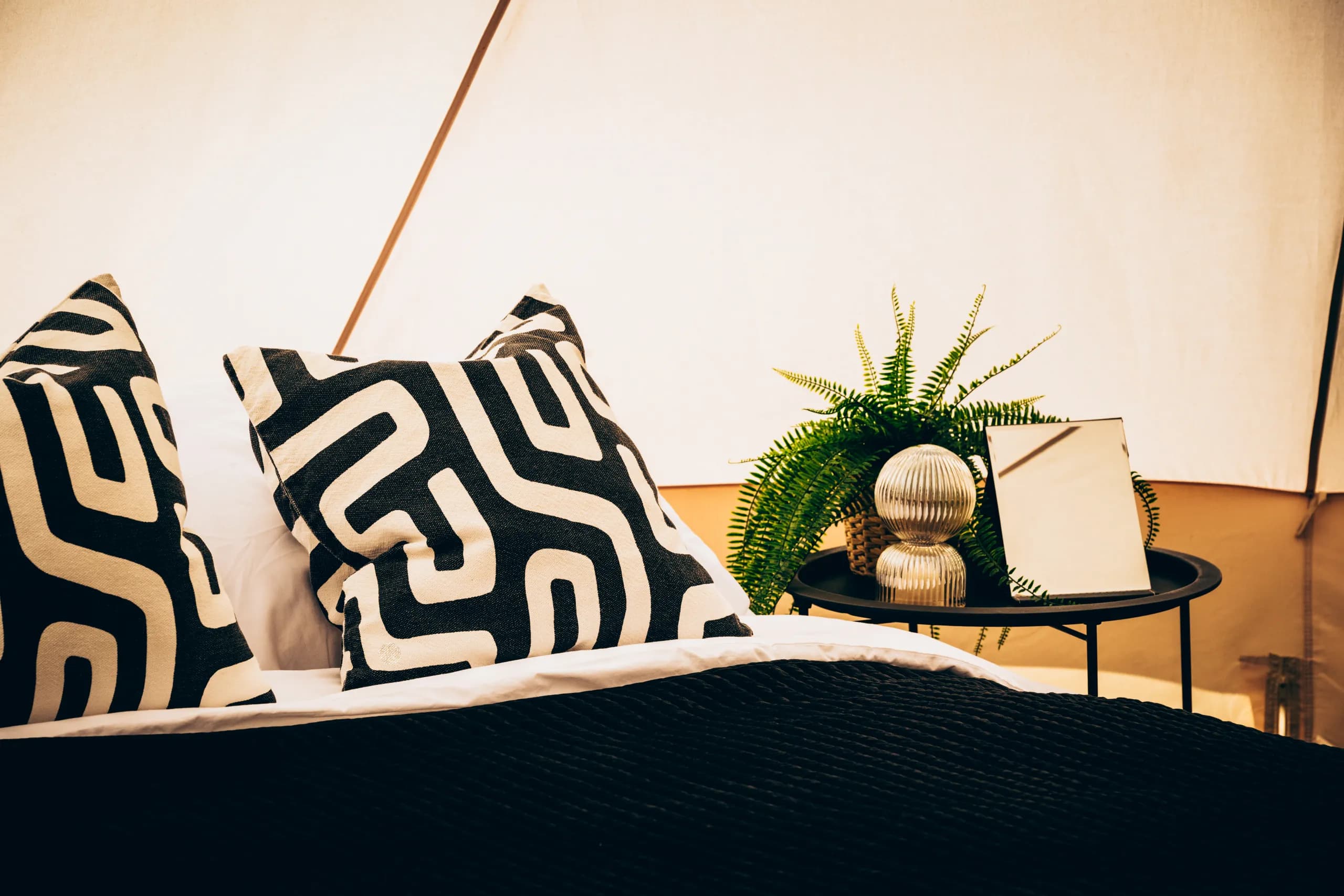 Close-up of black and white patterned cushions on white linen, with a fern and vanity mirror on the bedside table, inside a Hotel Melody Encore Lounge bell tent.