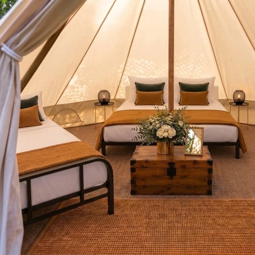 Two double beds dressed in white linen with green and mustard cushions inside a Hotel Melody Studio Lounge bell tent, with a wooden trunk, flowers and lanterns.