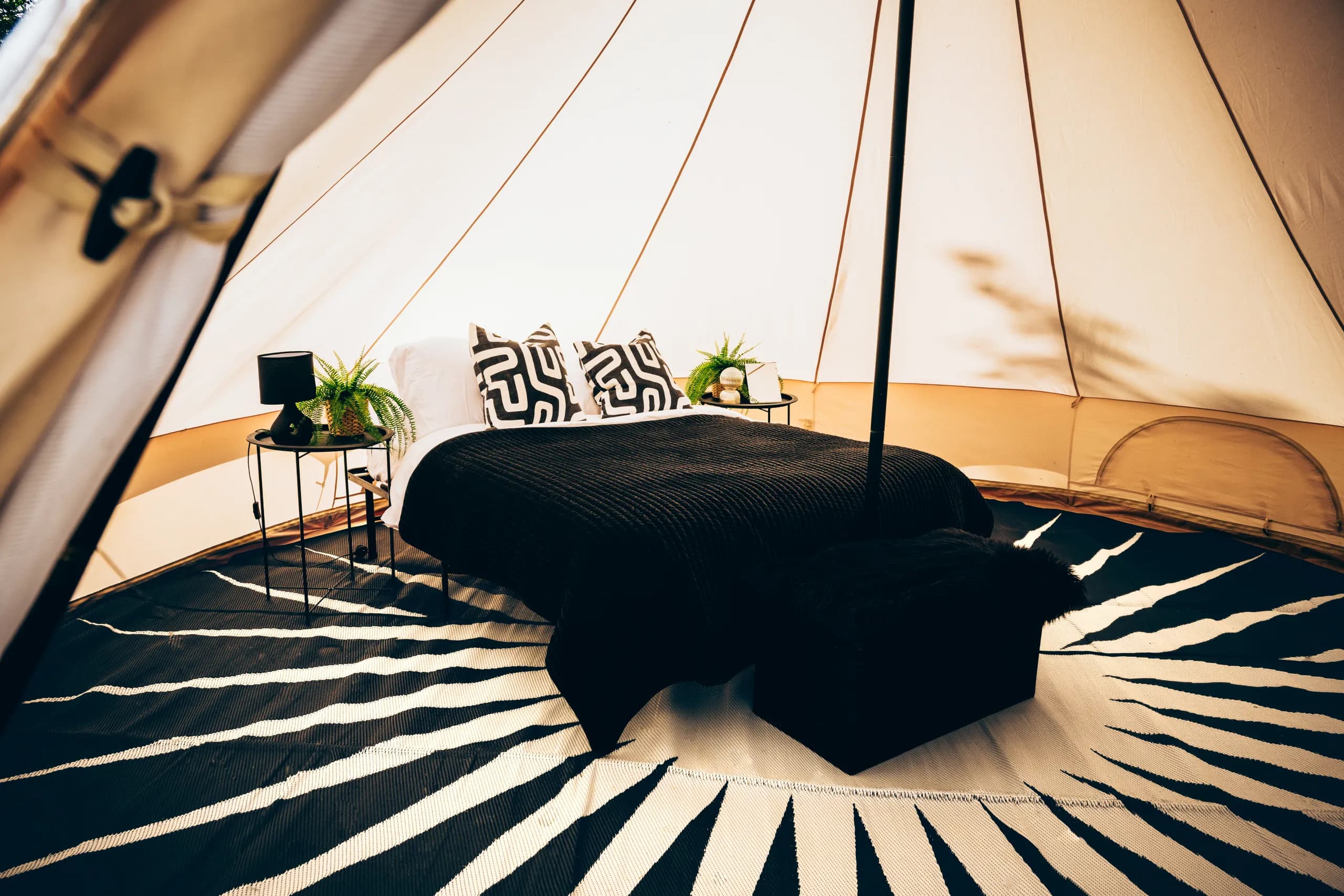 Looking through the entrance of a Hotel Melody Encore Lounge bell tent at a double bed with a black throw, bedside tables and a black and white sunburst floor rug.