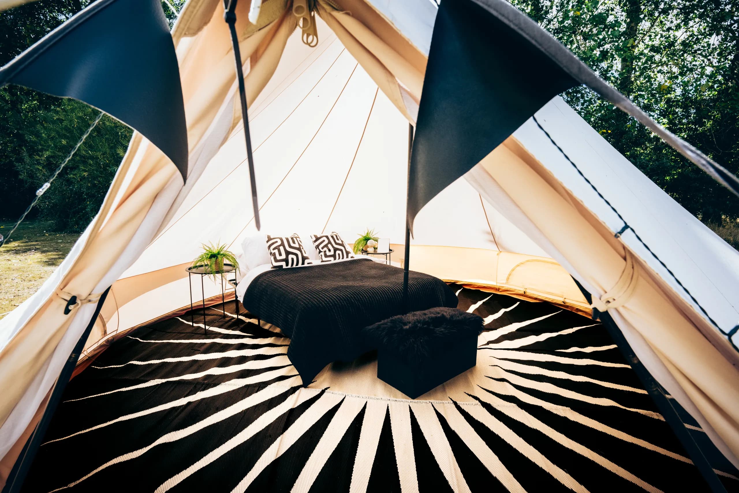 A double bed with a black throw and footstool on a sunburst rug, framed by black bunting at the entrance of a Hotel Melody Encore Lounge bell tent.