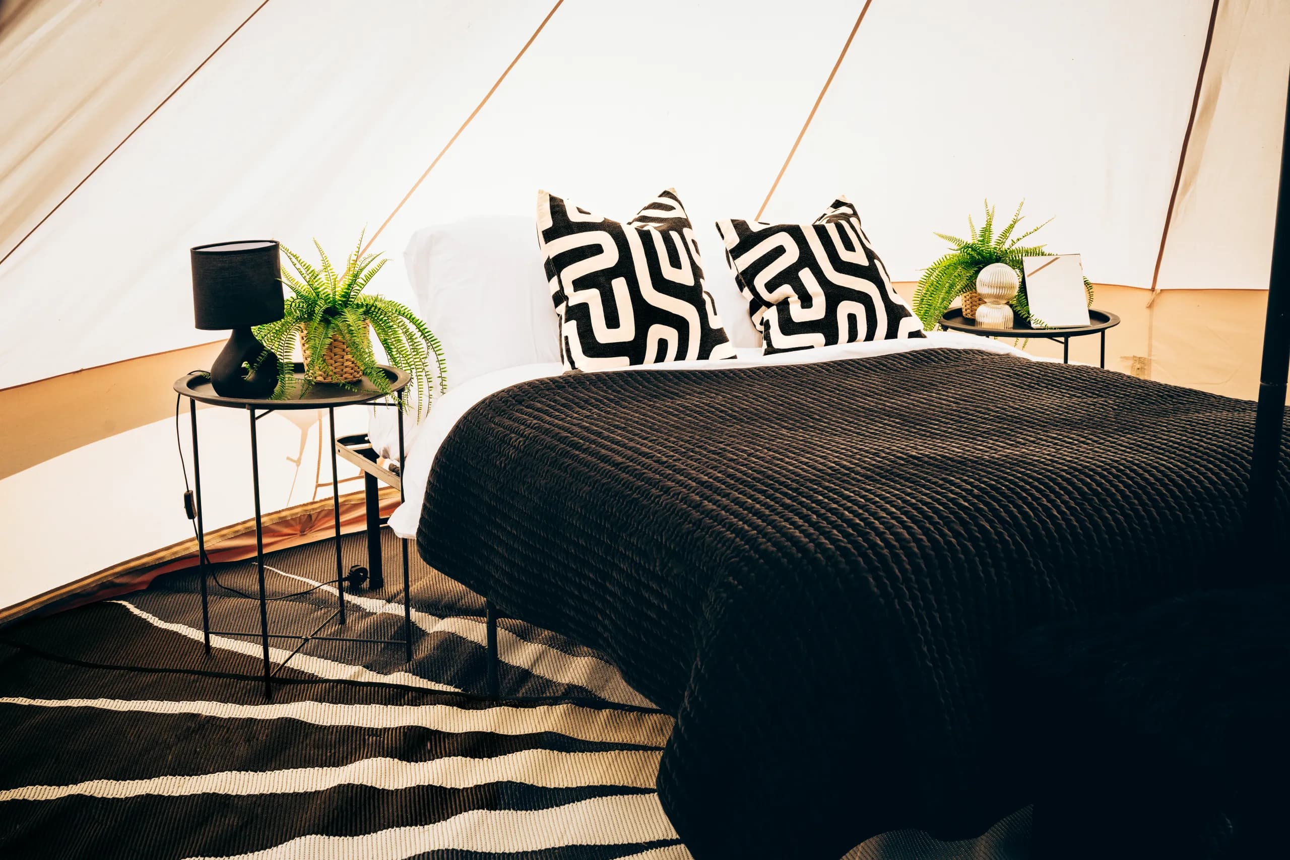 A double bed dressed with a black throw and black and white cushions between two bedside tables with a lamp and ferns, inside a Hotel Melody Encore Lounge bell tent.