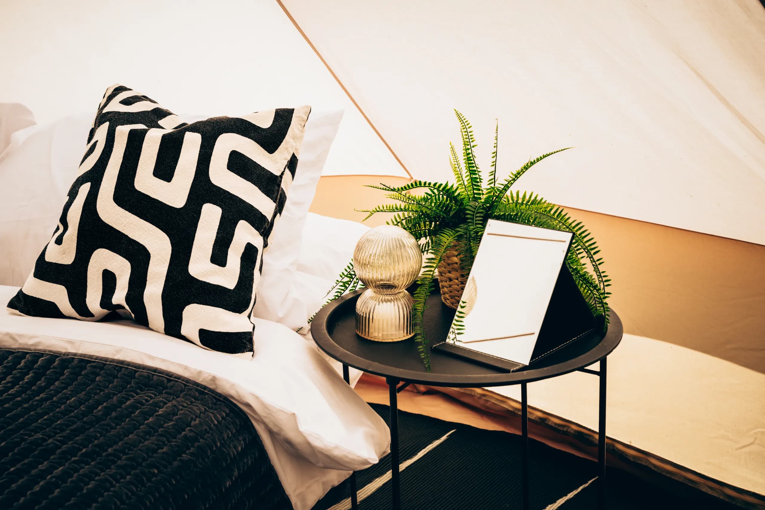 A black and white patterned cushion on a made-up bed beside a round side table with a fern, gold vase and vanity mirror, inside a Hotel Melody Encore Lounge bell tent.
