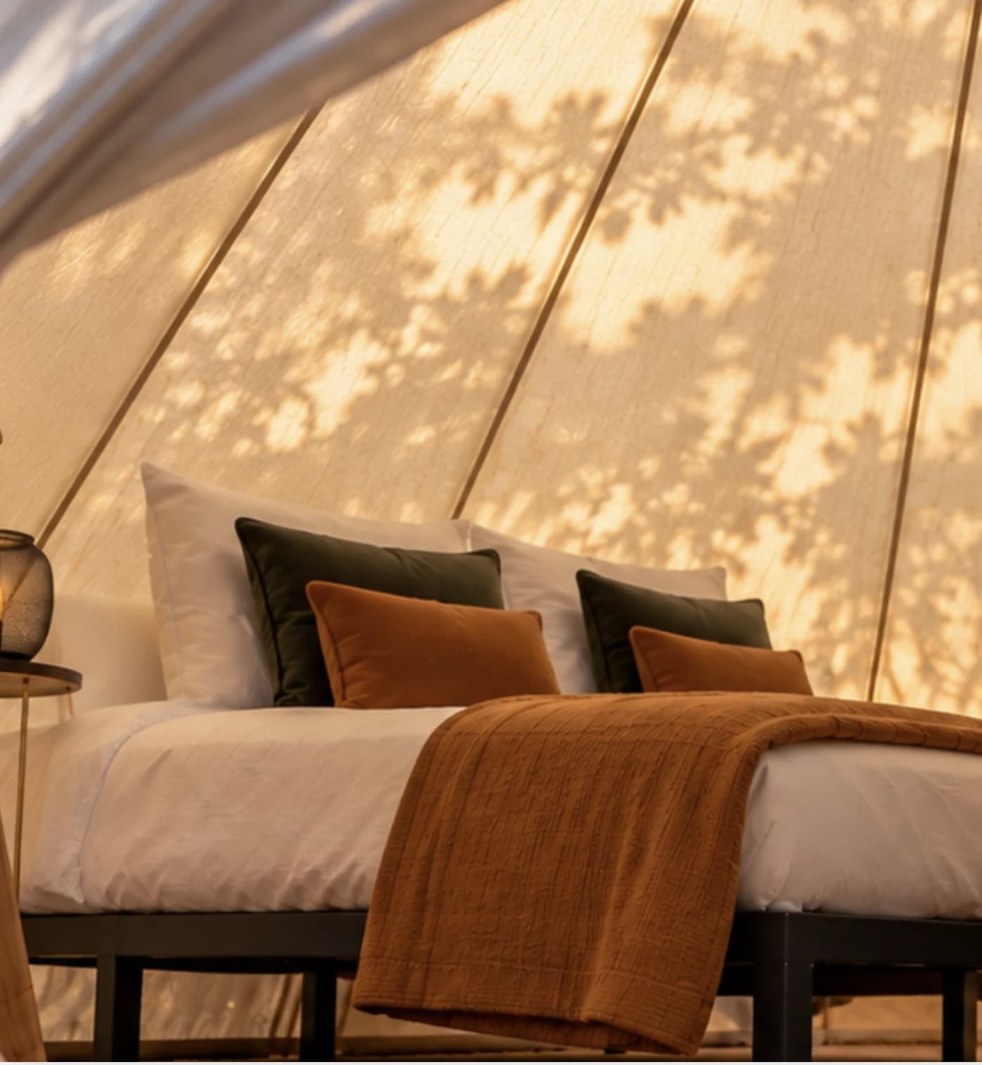 A double bed with green and burnt-orange cushions and a woven throw, under dappled light on the canvas walls of a Hotel Melody Studio Lounge bell tent.